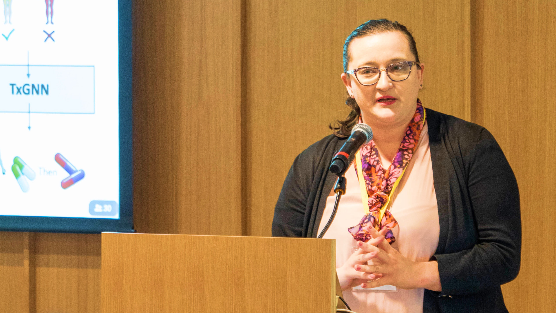 Marinka Zitnik, a woman wearing glasses and a colorful scarf, speaks into a microphone while standing at a podium, with a presentation slide displaying medical graphics in the background.
