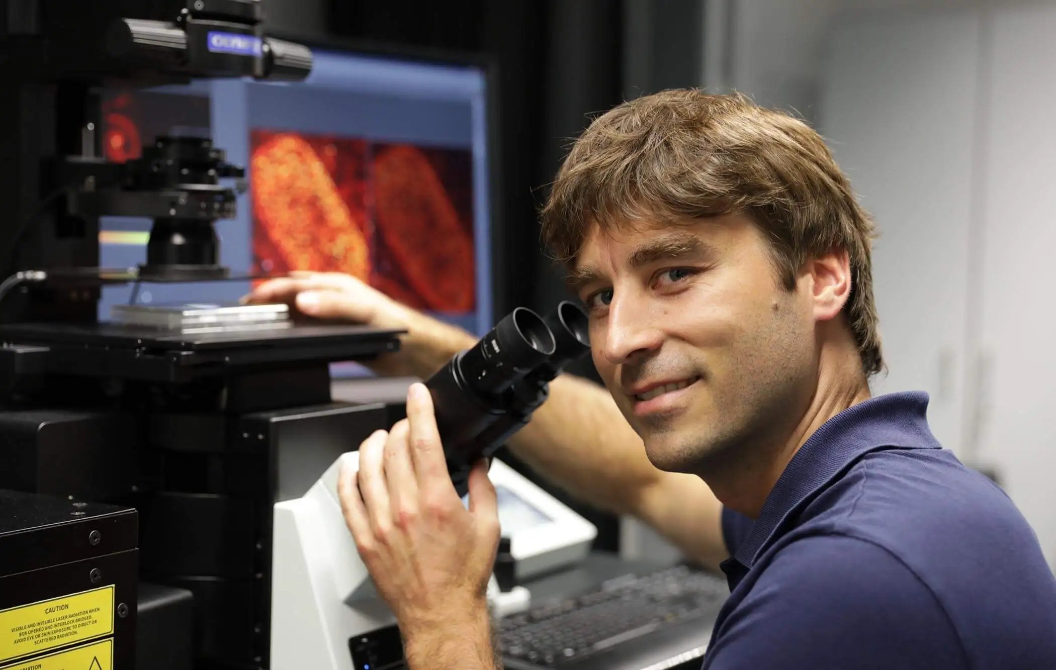 A man works at a microscope. Pictures of cells are in the background.
