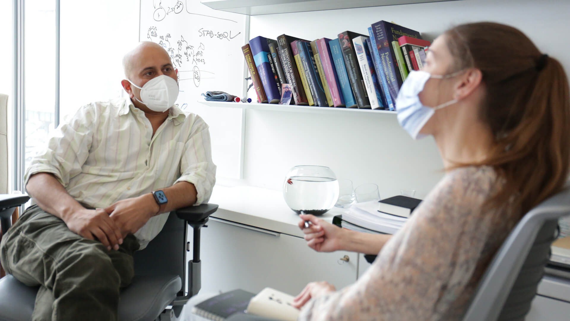 A man and woman wearing masks sit in an office chatting.